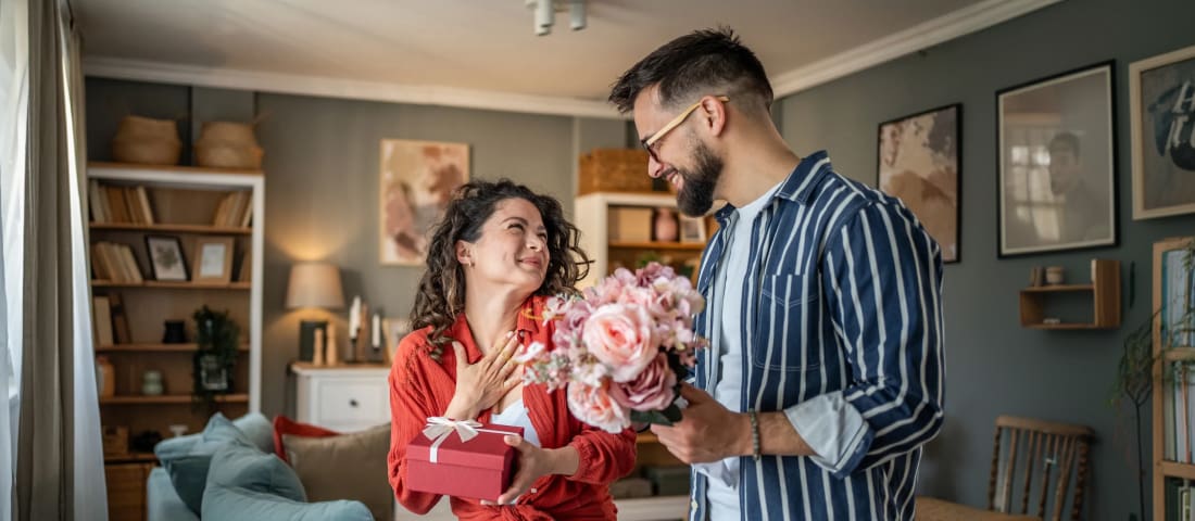 Pareja joven en una sala de estar, la mujer sostiene un regalo con la mano derecha mientras el hombre le da un ramo de flores.