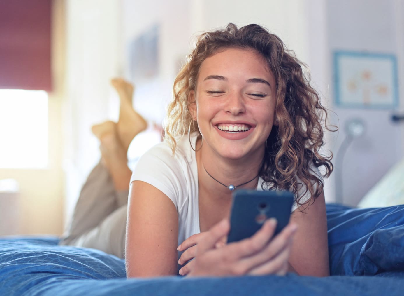 Una mujer sonriendo, viendo su teléfono mientras está acostada en una cama.