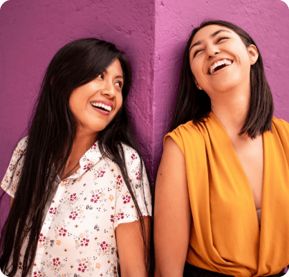 Dos mujeres sonrientes apoyadas en una pared morada en la calle.
